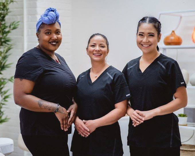 Three smiling women standing together wearing professional in a spa or clinic setting.