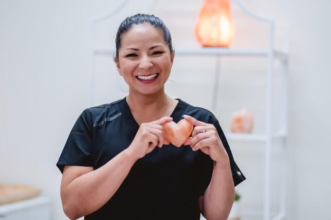 Mud Mask Facials at Zen'd Out Massage Spa A therapist smling towards the camera while holding a heart stone